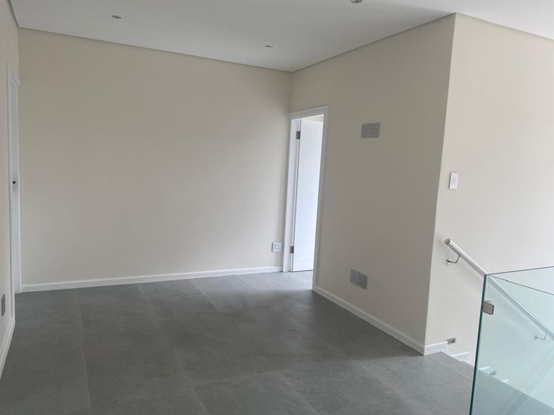 A bright, modern hallway featuring neutral walls, large format grey floor tiles, and a contemporary glass balustrade with a metal handrail.