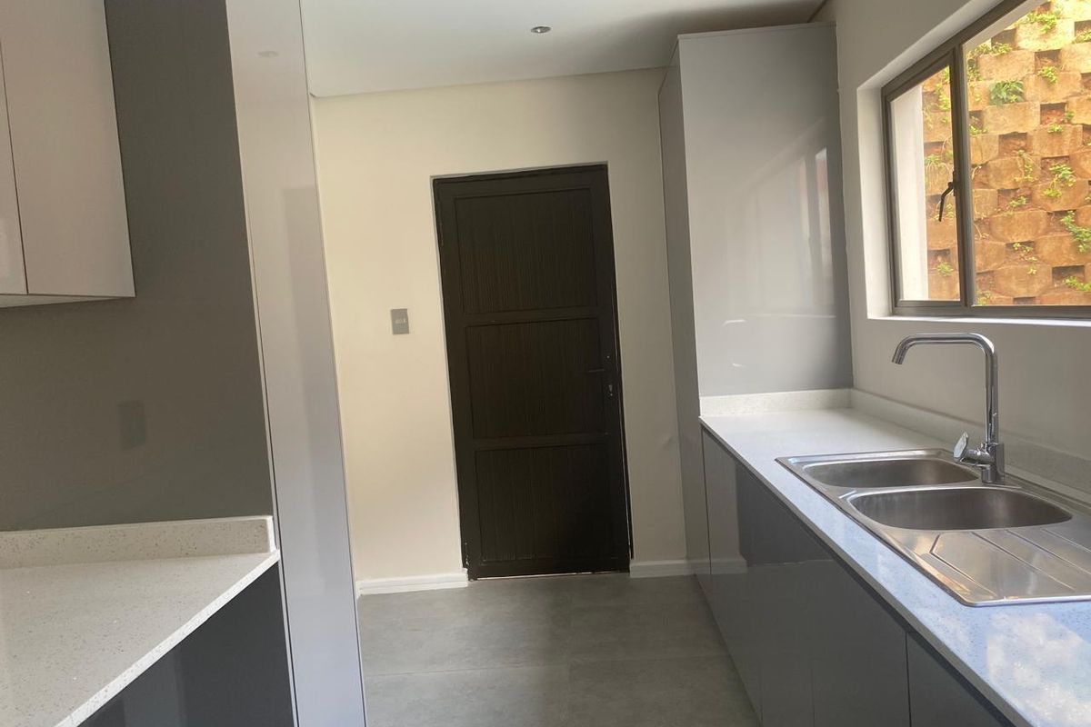 A modern, clean kitchen featuring sleek grey cabinetry, stone countertops, and a stainless steel double sink with a view of the exterior retaining wall.