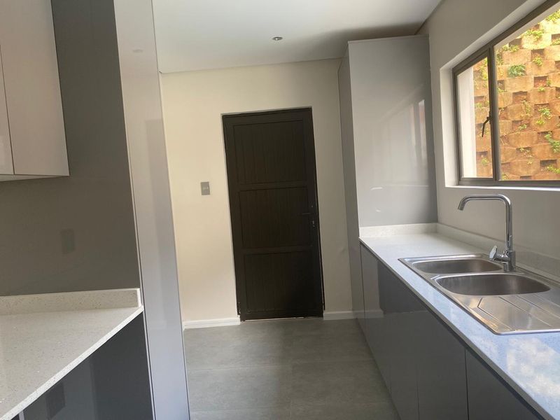 A modern, clean kitchen featuring sleek grey cabinetry, stone countertops, and a stainless steel double sink with a view of the exterior retaining wall.