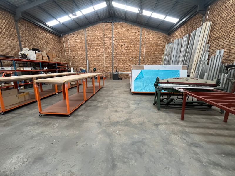 Spacious industrial workshop featuring exposed brick walls, high steel truss ceilings with skylights, and durable concrete flooring, suitable for manufacturing or storage operations.