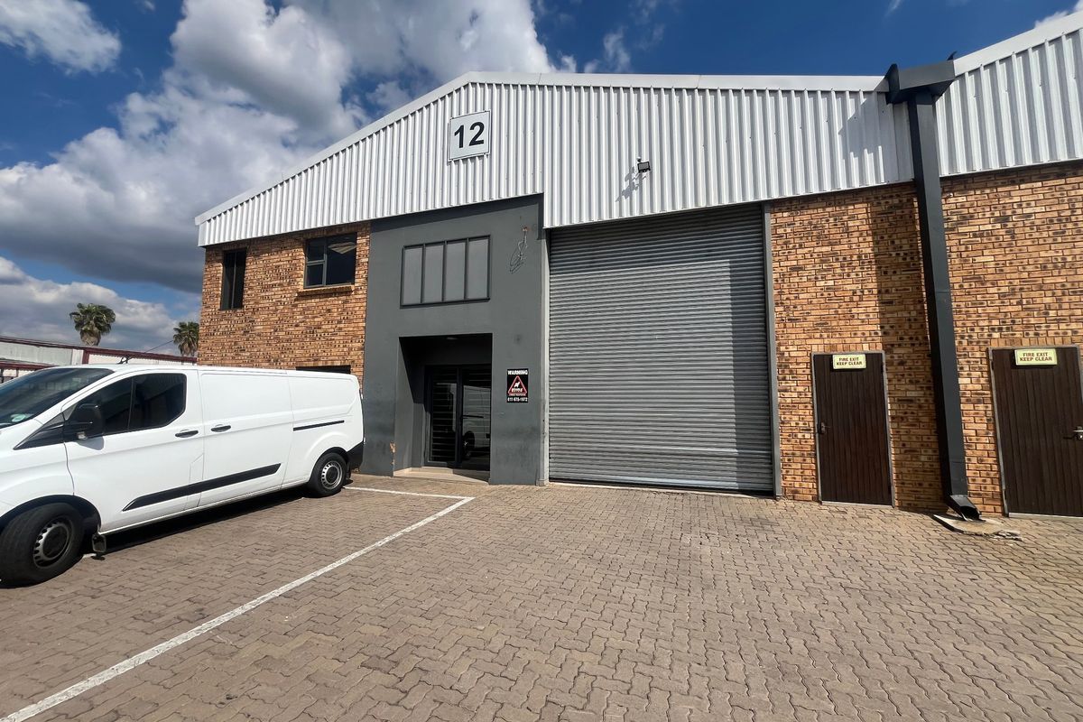 Front elevation of an industrial unit featuring a large roller shutter door, brick and metal cladding construction, and dedicated paved parking area.