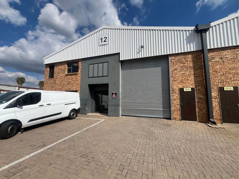Front elevation of an industrial unit featuring a large roller shutter door, brick and metal cladding construction, and dedicated paved parking area.