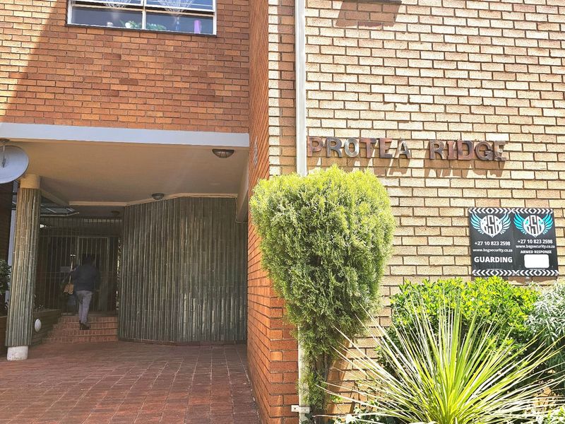 The front entrance of the Protea Ridge apartment complex, featuring a brick facade, paved walkway, and secure gated access.