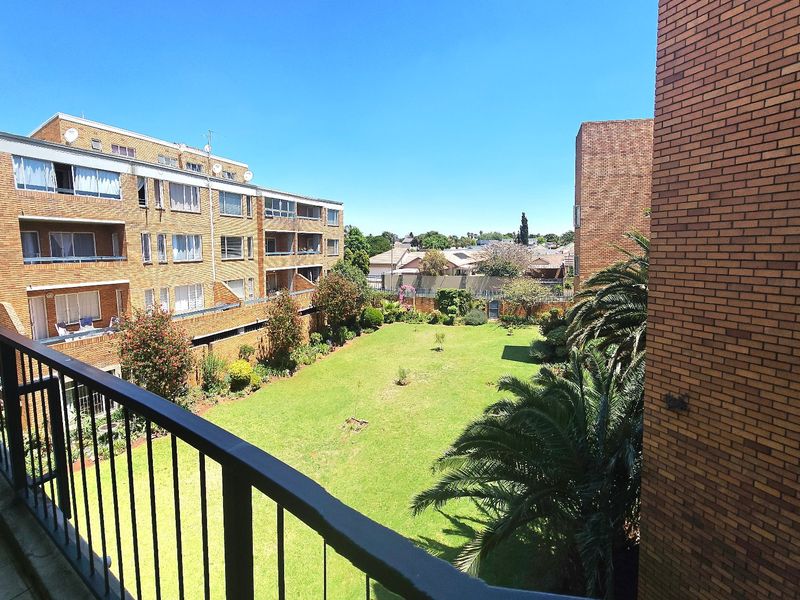 A spacious balcony with a metal balustrade overlooking a well-maintained communal garden and surrounding apartment complex.