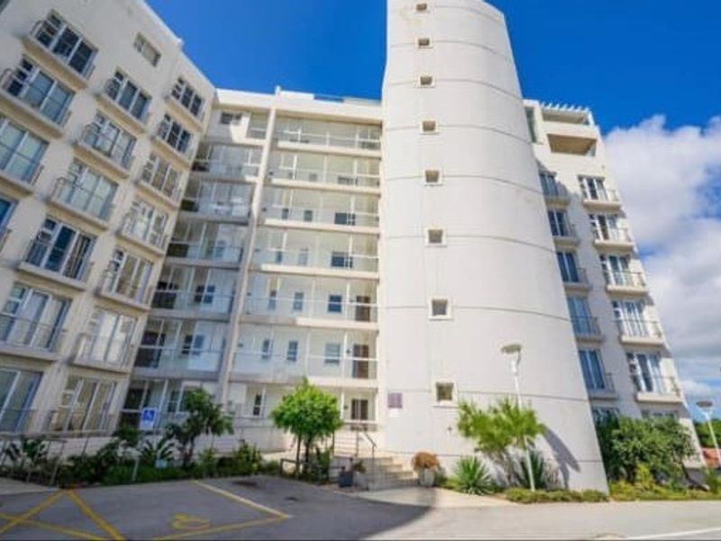 A multi-story apartment building featuring a prominent cylindrical architectural element, multiple balconies with glass balustrades, and a paved front entrance area.