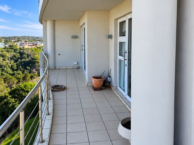 A spacious covered balcony featuring tiled flooring, stainless steel balustrades, and glass sliding door access, offering scenic elevated views of the surrounding landscape.