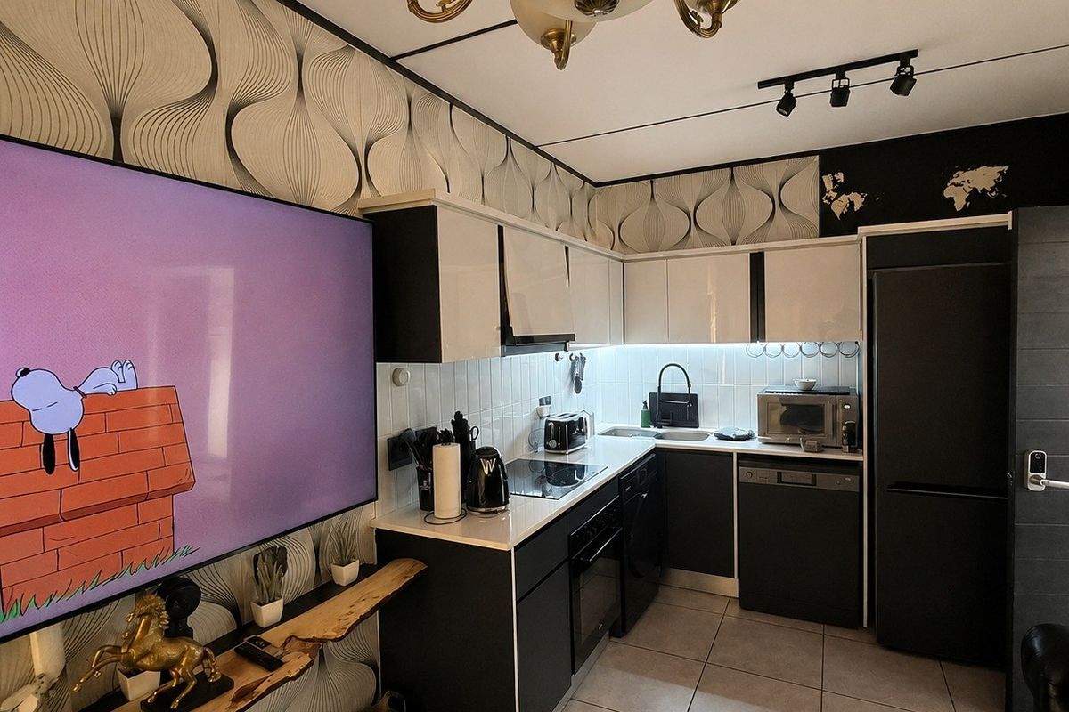 A modern, compact kitchen featuring high-contrast black and white cabinetry, integrated appliances, tiled flooring, and a decorative patterned wallpaper finish.
