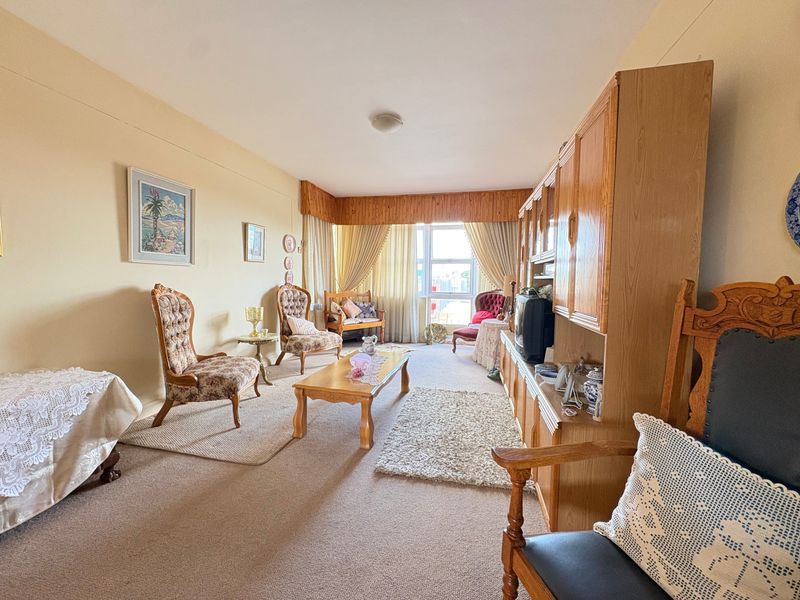 A spacious living room featuring carpeted flooring and a large window with a wooden pelmet, offering a dated but functional living space.