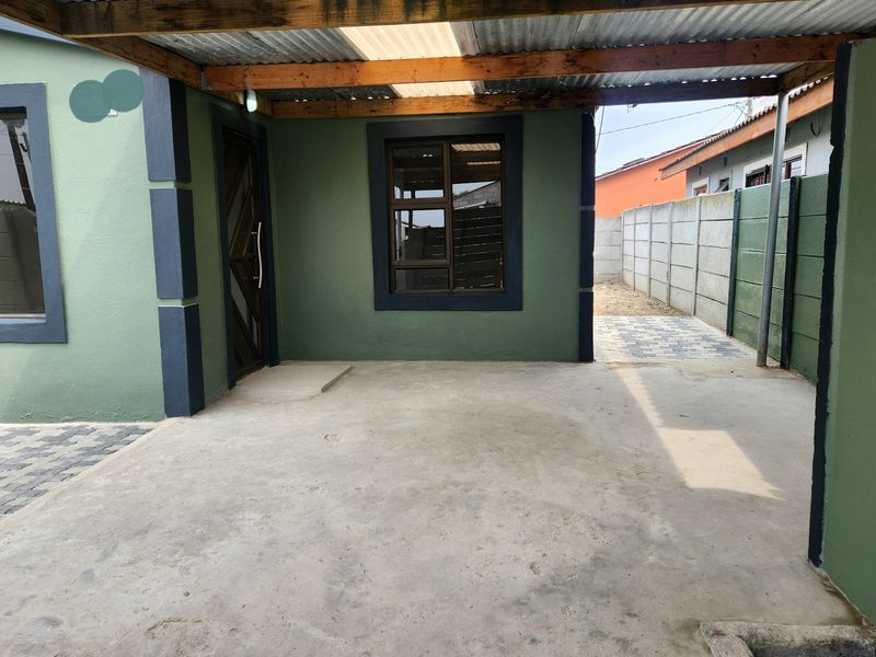 A covered outdoor patio area featuring a concrete floor, timber-framed corrugated roof, and access to a paved side yard with precast boundary walls.