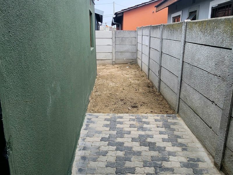A narrow side or rear yard area featuring a paved walkway and precast concrete boundary walling, with an unlandscaped soil section.