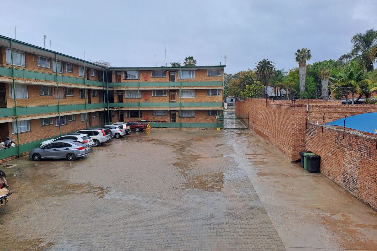 An exterior view of an apartment complex courtyard featuring a paved parking area, brick building facades, and a secure perimeter wall with electric fencing.