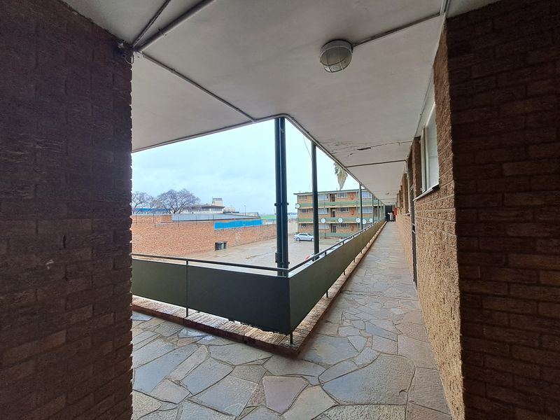 An open-air apartment complex walkway featuring exposed brick walls, stone paving, and a metal balustrade overlooking the parking area.