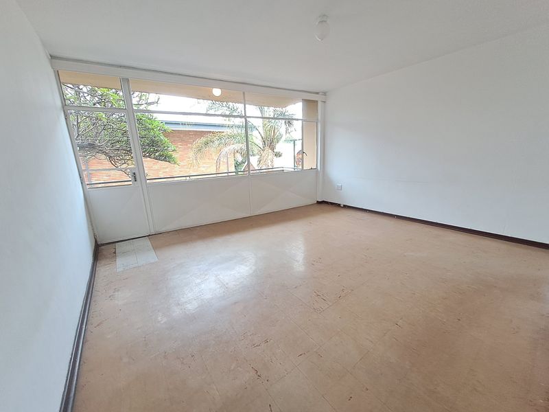 An empty living room featuring large glass doors leading to a balcony, with neutral walls and worn vinyl flooring throughout.