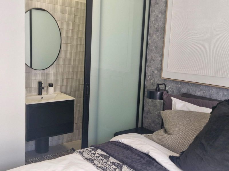 A modern bedroom featuring a sleek wall-mounted vanity with a basin and round mirror, separated from the sleeping area by a sliding glass door.