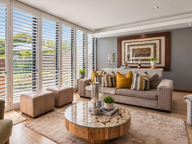 A bright and modern living room featuring timber flooring, recessed ceiling lighting, and large floor-to-ceiling windows with white plantation shutters.