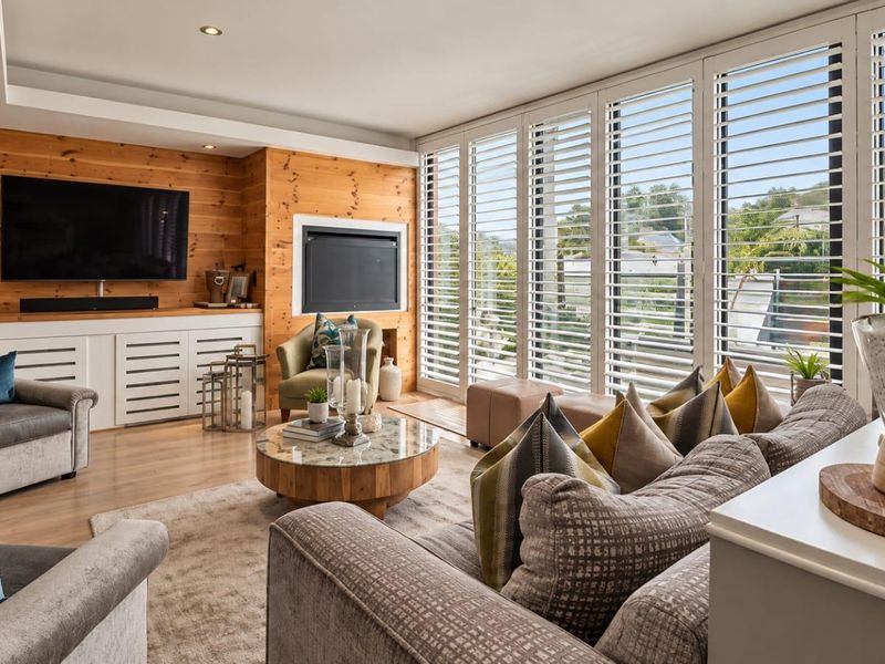 A well-appointed living room featuring timber wall cladding, built-in cabinetry, a fireplace, and large windows with plantation shutters providing ample natural light.