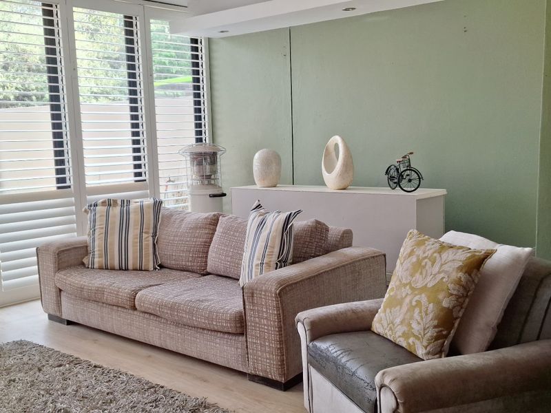 A bright living room featuring light laminate flooring, recessed downlights, and large windows fitted with white plantation shutters.
