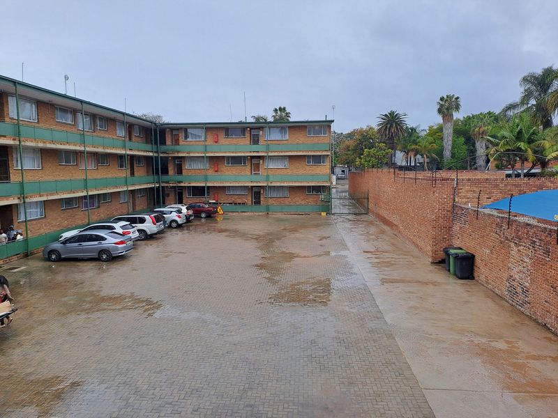 An exterior view of an apartment complex courtyard featuring a paved parking area, brick building facades, and a secure perimeter wall with electric fencing.