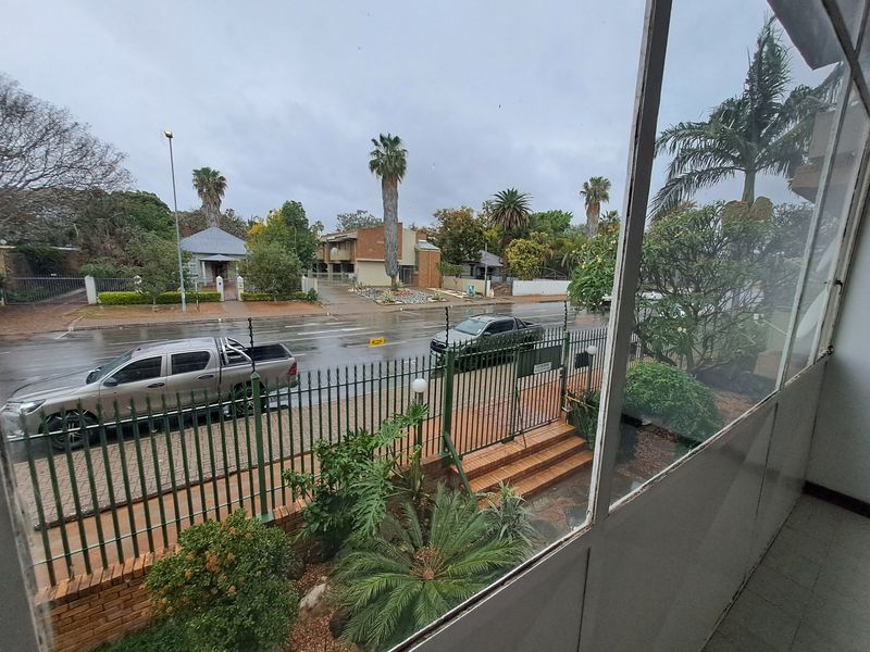 View from an elevated position overlooking a residential street, featuring metal security fencing, paved driveway, and established garden landscaping.