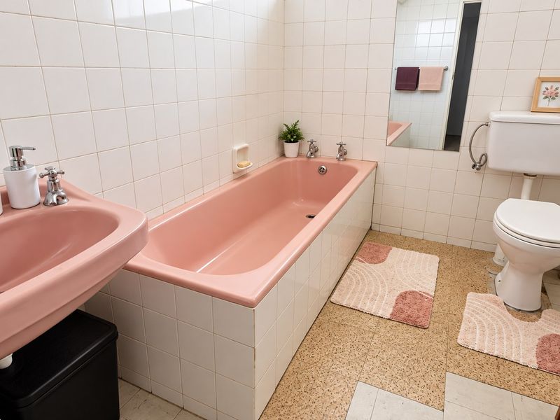 A dated bathroom featuring original pink bathtub and basin, white wall tiling, and terrazzo-style flooring, offering significant potential for a modern renovation.