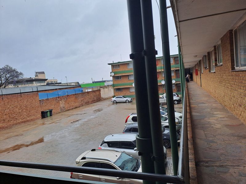 An exterior view from an apartment balcony overlooking a paved parking area and adjacent building facade.