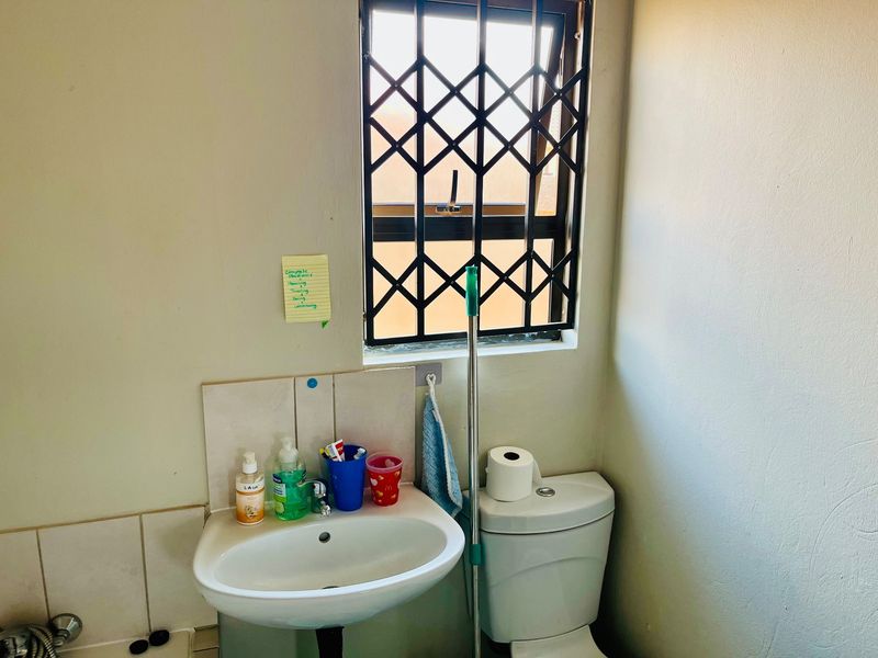 A functional bathroom featuring a pedestal basin with tiled splashback, a standard toilet, and a window fitted with a security gate.