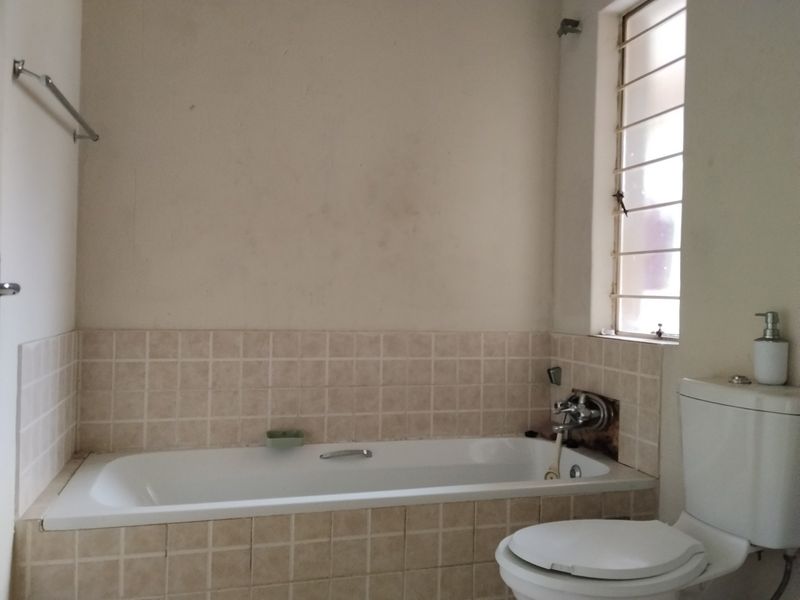 A dated bathroom featuring a built-in bathtub with tiled surround, a standard toilet, and a window with security bars.