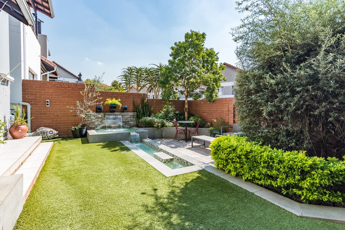 A well-maintained rear garden featuring a brick boundary wall, artificial lawn, timber decking, and a built-in water feature.