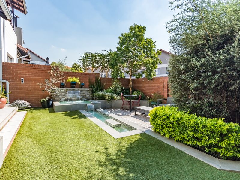 A well-maintained rear garden featuring a brick boundary wall, artificial lawn, timber decking, and a built-in water feature.