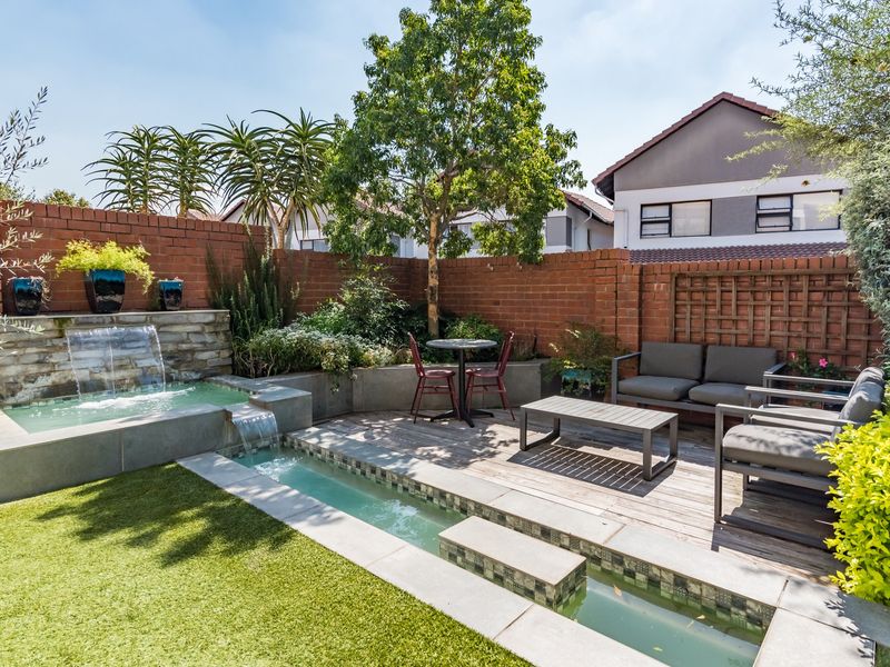 A landscaped garden featuring a tiered water feature, timber decking, artificial lawn, and brick boundary walls, creating a tranquil outdoor living space.