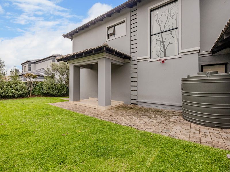 Rear exterior view featuring a covered patio, paved area, water storage tank, and well-maintained lawn against a modern grey facade.