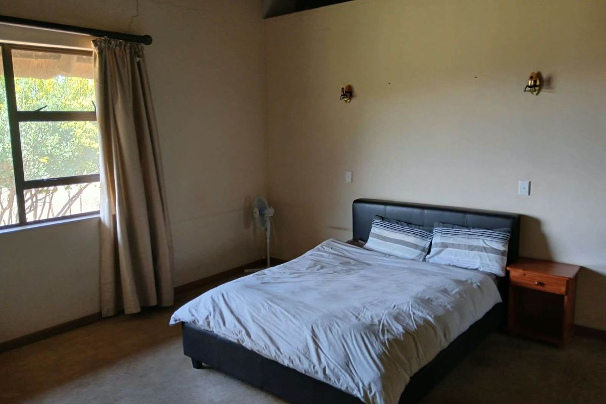 A bedroom featuring exposed timber ceiling beams, wall-mounted lighting, and a large window. The space shows signs of wear including visible wall cracks.