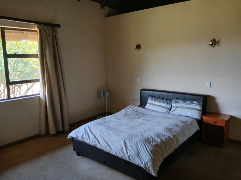 A bedroom featuring exposed timber ceiling beams, wall-mounted lighting, and a large window. The space shows signs of wear including visible wall cracks.