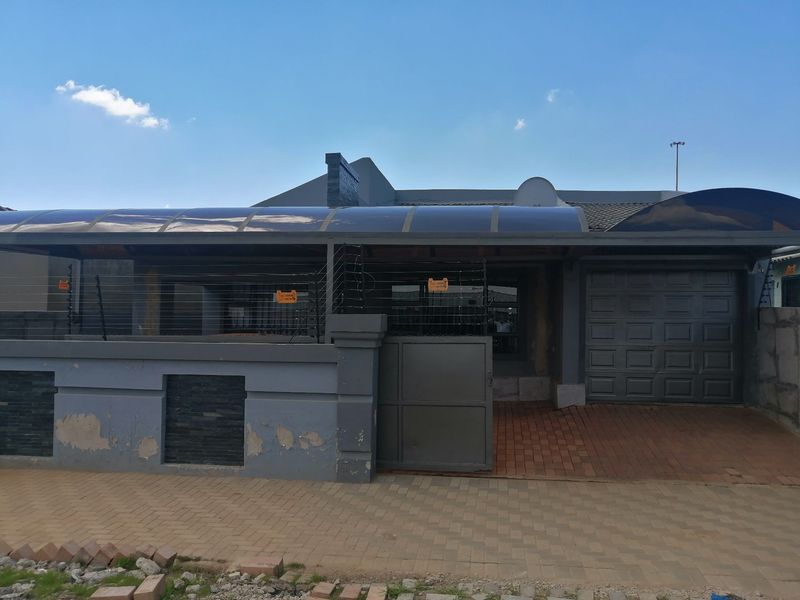 Front elevation of the property featuring a paved driveway, sectional garage door, covered carport area, and perimeter boundary wall with electric fencing.