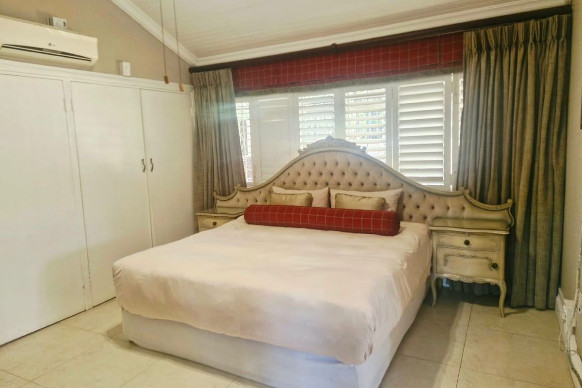 A spacious bedroom featuring vaulted ceilings, tiled flooring, built-in cupboards, and a wall-mounted air conditioning unit with plantation shutters for light control.
