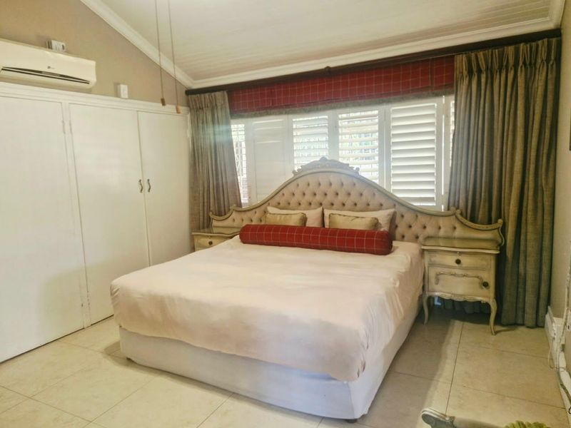 A spacious bedroom featuring vaulted ceilings, tiled flooring, built-in cupboards, and a wall-mounted air conditioning unit with plantation shutters for light control.