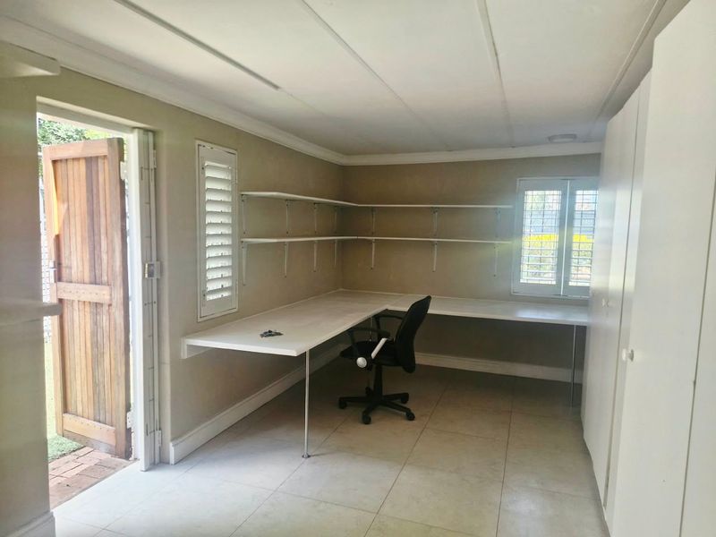 A functional home office featuring a built-in L-shaped desk, wall-mounted shelving, tiled floors, and direct access to the exterior via a timber door.