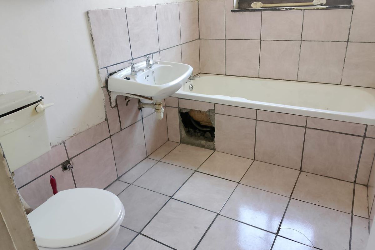 A dated bathroom featuring a bathtub, wall-mounted basin, and toilet, requiring repairs to the tiled bathtub panel and general cosmetic updates.