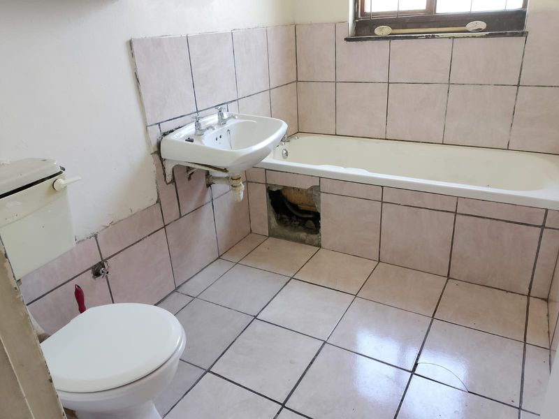 A dated bathroom featuring a bathtub, wall-mounted basin, and toilet, requiring repairs to the tiled bathtub panel and general cosmetic updates.