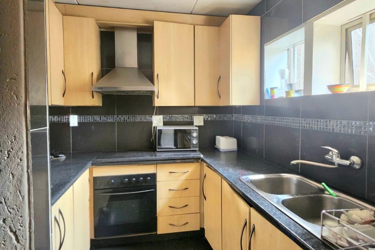 A functional galley-style kitchen featuring light wood cabinetry, dark granite countertops, a built-in oven, extractor fan, and a double stainless steel sink.