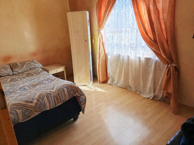A bedroom featuring laminate flooring and a window with curtains, showing significant wear to the wall finishes.