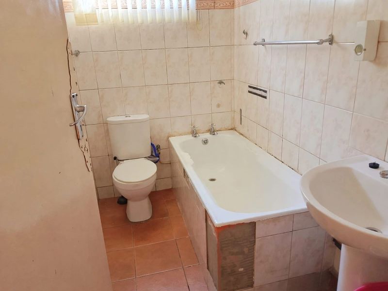 A dated bathroom featuring a standard bathtub, pedestal sink, and toilet, requiring renovation and repair to the wall tiles and bathtub paneling.