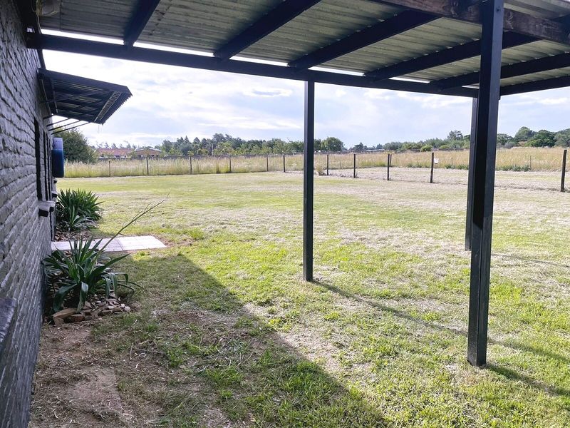 A covered outdoor patio area featuring a corrugated metal roof and steel support posts, overlooking a large, open grass garden.