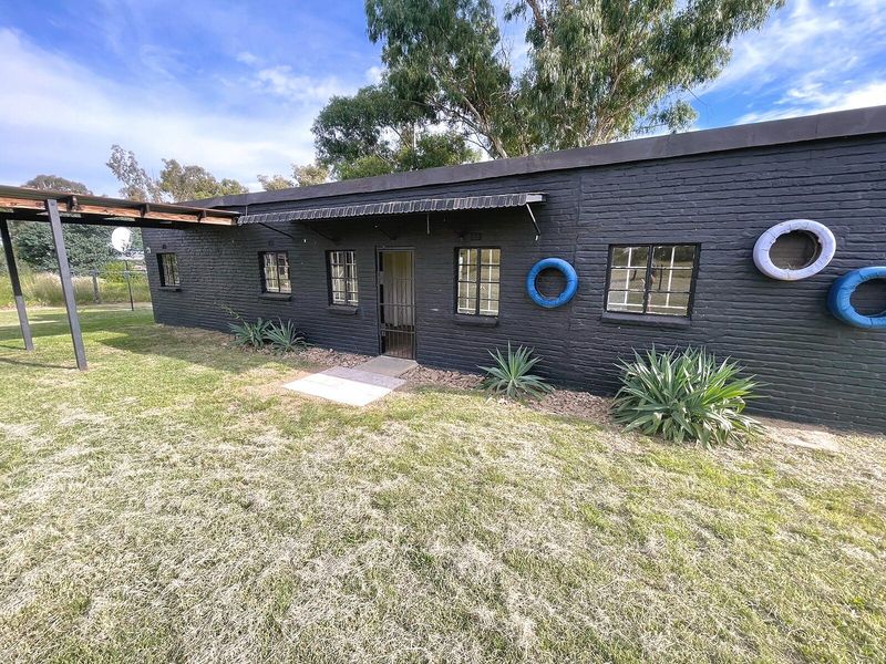 Rear view of a single-story house featuring a dark painted brick facade, steel-framed windows, and an attached covered patio area overlooking the garden.
