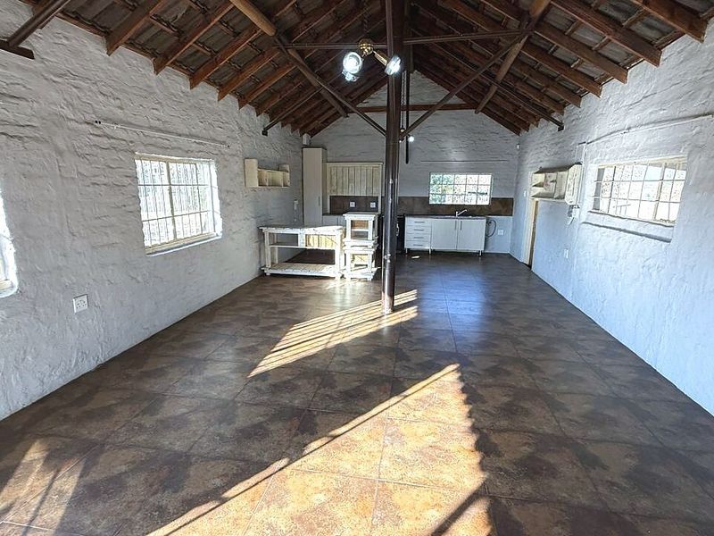 Spacious open-plan living area featuring exposed timber roof trusses, textured masonry walls, tiled flooring, and a functional kitchen corner with built-in cabinetry.