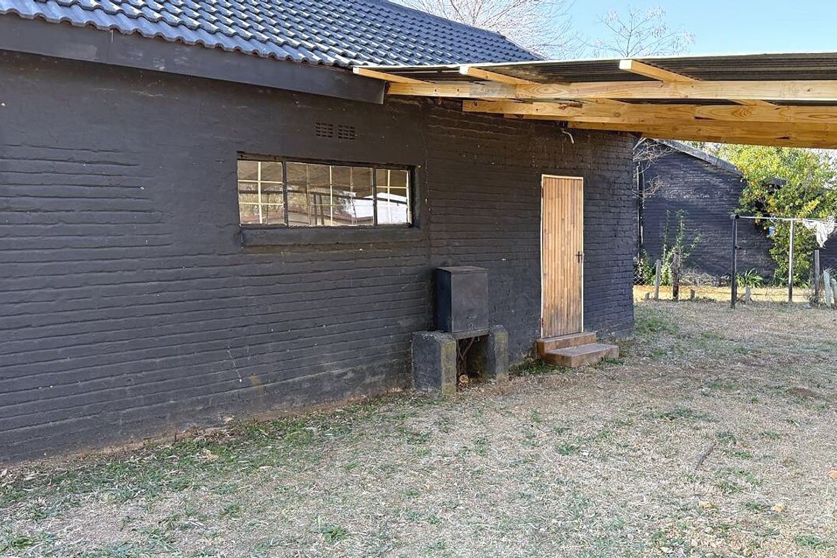 Rear exterior view of a dark-painted brick house featuring a tiled roof, a built-in outdoor braai, and a simple corrugated iron lean-to structure.