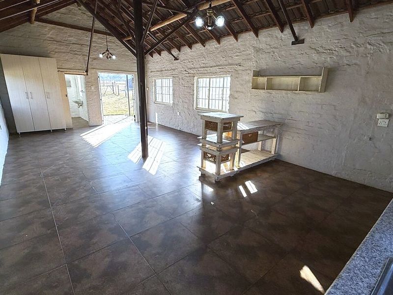 Spacious open-plan area featuring rustic painted stone walls, exposed timber roof trusses, and tiled flooring, offering a versatile layout with integrated kitchen elements.