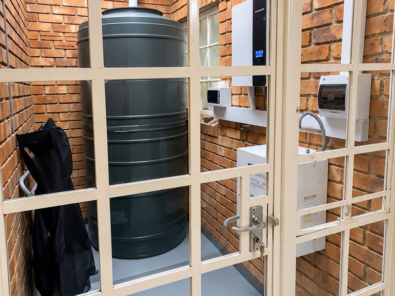 A clean utility space featuring a water storage tank, inverter system, and electrical distribution board, enclosed by a secure glass-paneled door.