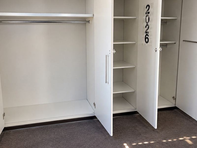 A clean bedroom interior featuring a large white built-in wardrobe with shelving and hanging space, complemented by neutral carpet flooring.