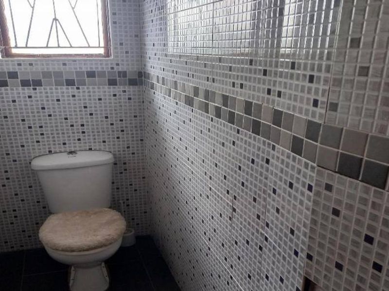A small bathroom featuring mosaic wall tiling and a standard toilet, with a window fitted with security bars.